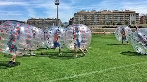 People wearing transparent inflatable bubble suits playing a soccer match on a sunny outdoor grass field.