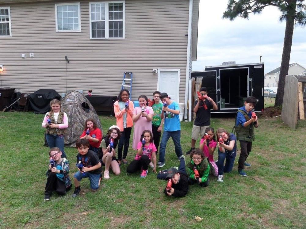 A large group of children poses outdoors in a backyard, holding toy blasters while facing the camera.