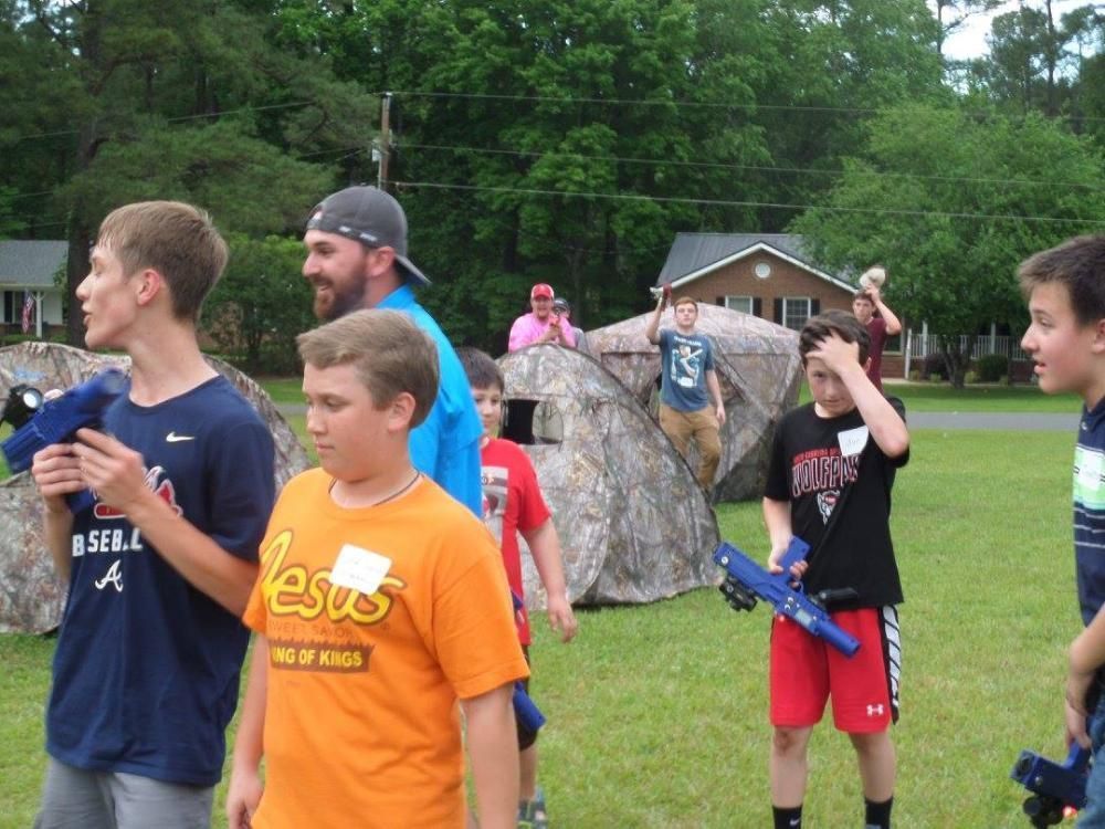 A group of people holding toy blasters stand near camouflage play tents on a grassy lawn with houses in the background.