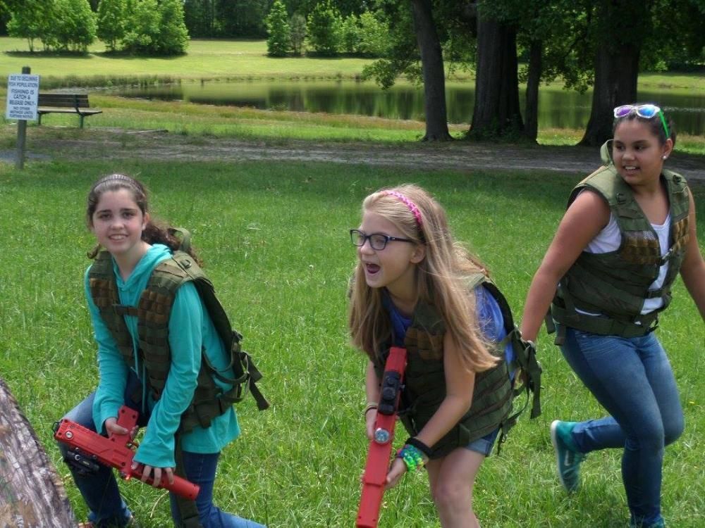 Three children wearing tactical vests and holding red toy blasters run across a grassy field near a pond.