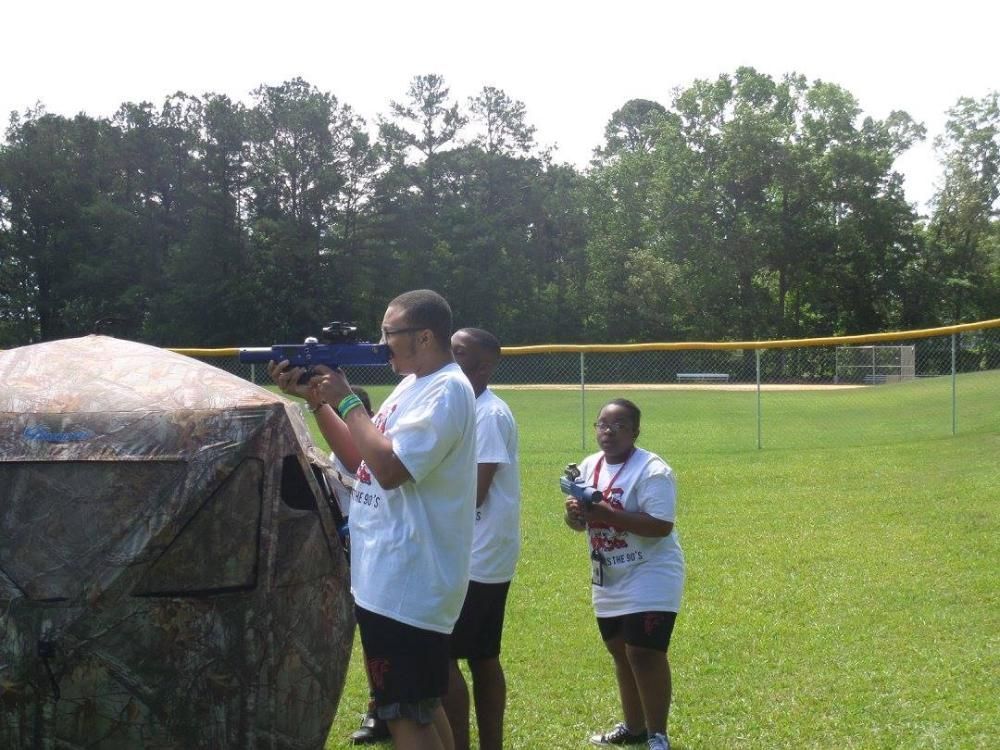 Three people in white t-shirts practice target shooting with toy blasters near a camouflage tent on a grassy field.