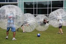 Four people wearing large, clear inflatable bubbles play soccer on a green lawn in front of a modern glass building.