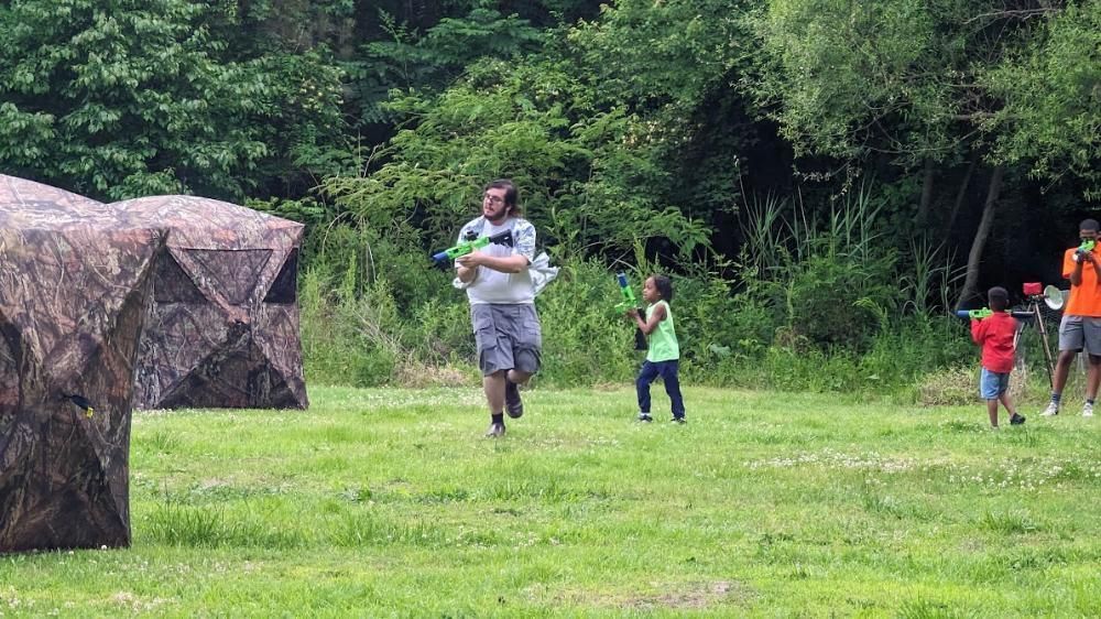 A group of people playing with toy blasters in a grassy field near camouflage-patterned inflatable obstacles.