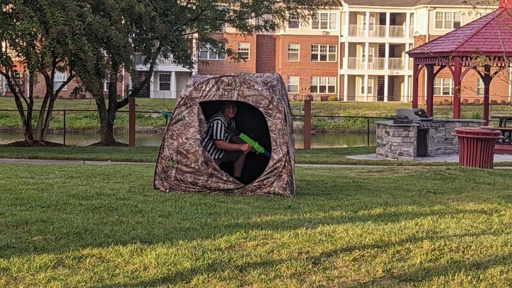 A person in a camouflage hunting blind holding a green toy water gun in a park with a gazebo in the background.