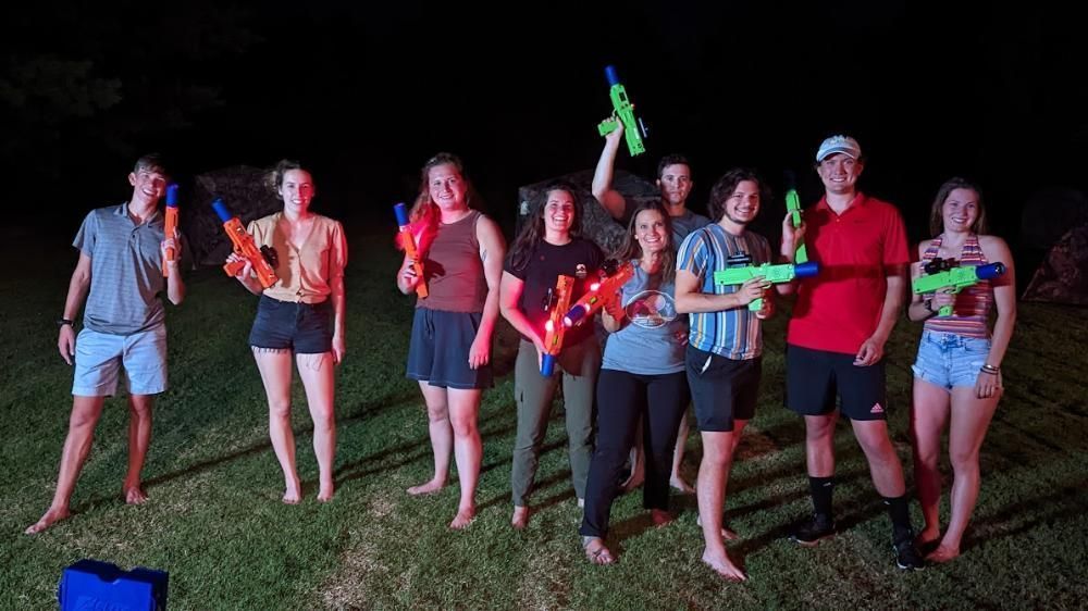 A group of nine people stand on a grassy field at night, smiling and holding neon-colored water guns.