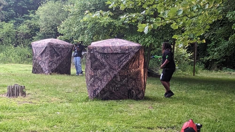 Two camouflage-patterned pop-up ground blinds stand in a grassy, wooded clearing with two people nearby.