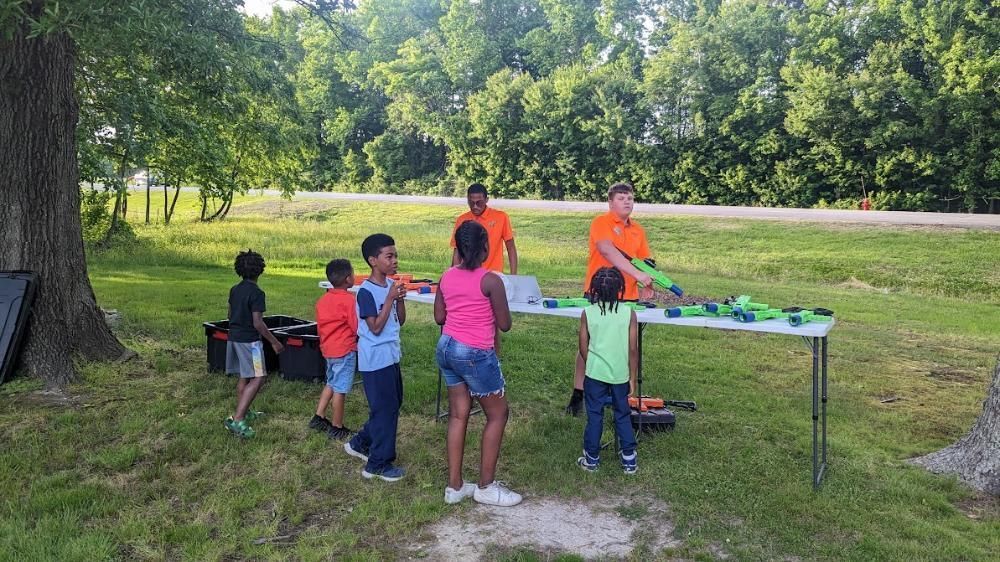 A group of children and two adults gather around a folding table outside, preparing to play with toy foam blasters.