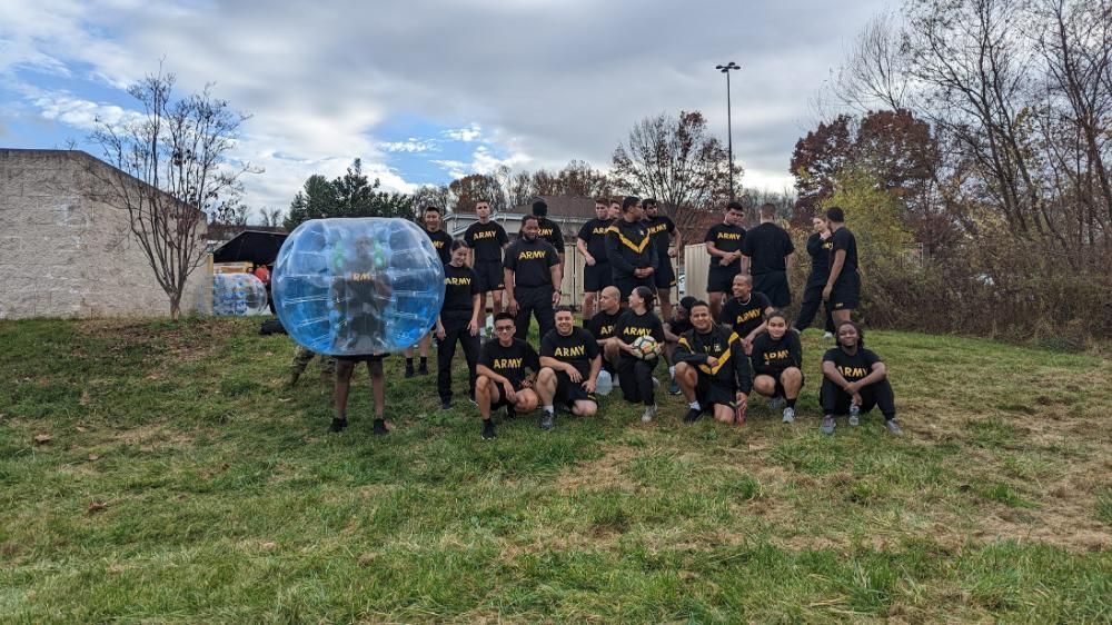 A group in athletic gear poses on a grassy field, with one person standing inside a large, clear bubble suit.