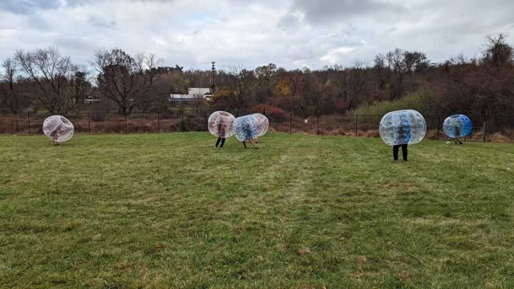 People in large, clear inflatable bubbles play in a grassy field under a cloudy sky.