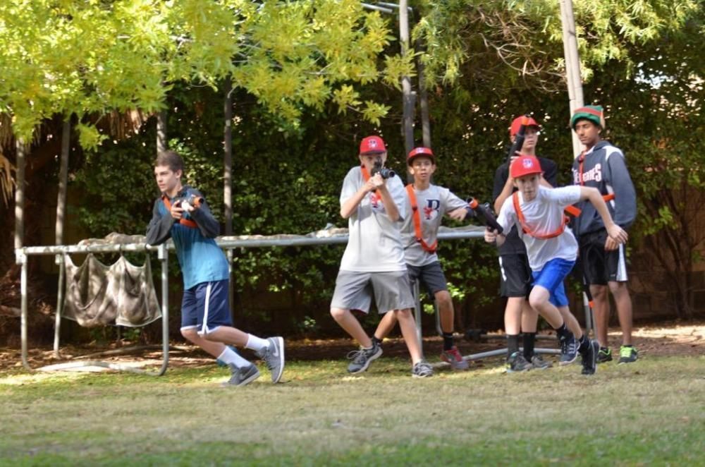 A group of people wearing game vests and holding toy guns running across a grassy field near trees and a trampoline.