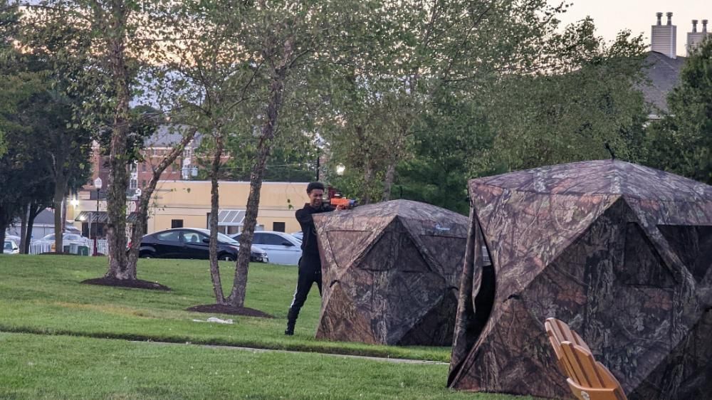 A person stands on a grassy field behind a camouflage tent, aiming an orange toy blaster in a park setting.