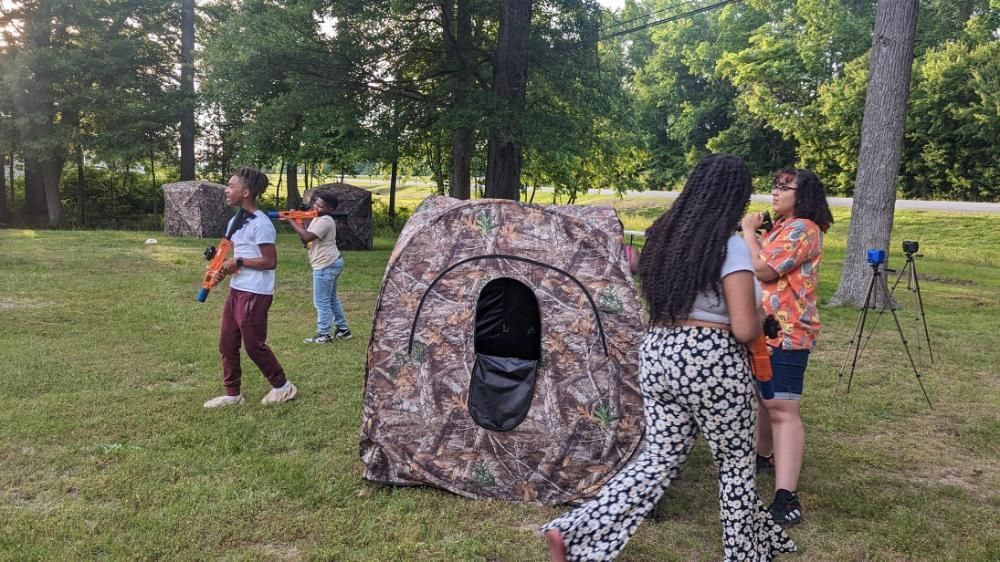 People play with toy blasters near camouflage ground blinds in a grassy, wooded outdoor area.