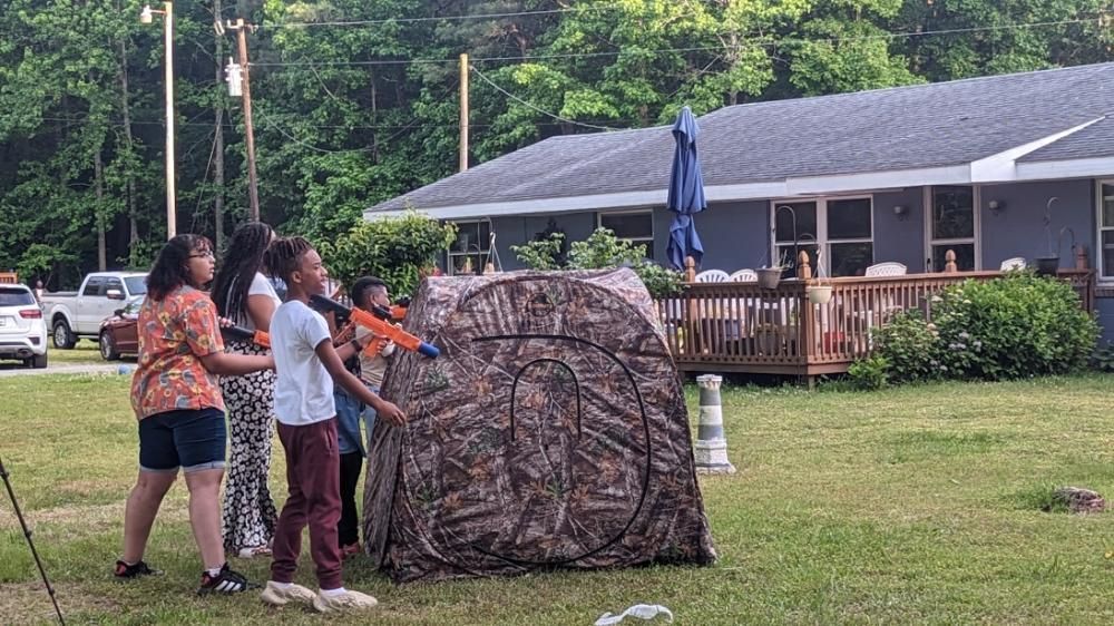 Four people stand in a grassy yard, aiming toy blasters at a camouflage hunting blind near a house with a wooden deck.
