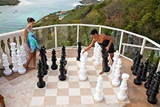 Two people play with oversized chess pieces on a balcony overlooking a bay.