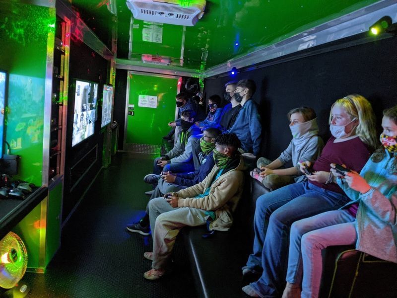 People wearing face masks sit on benches inside a neon-green mobile gaming trailer, playing video games on wall screens.