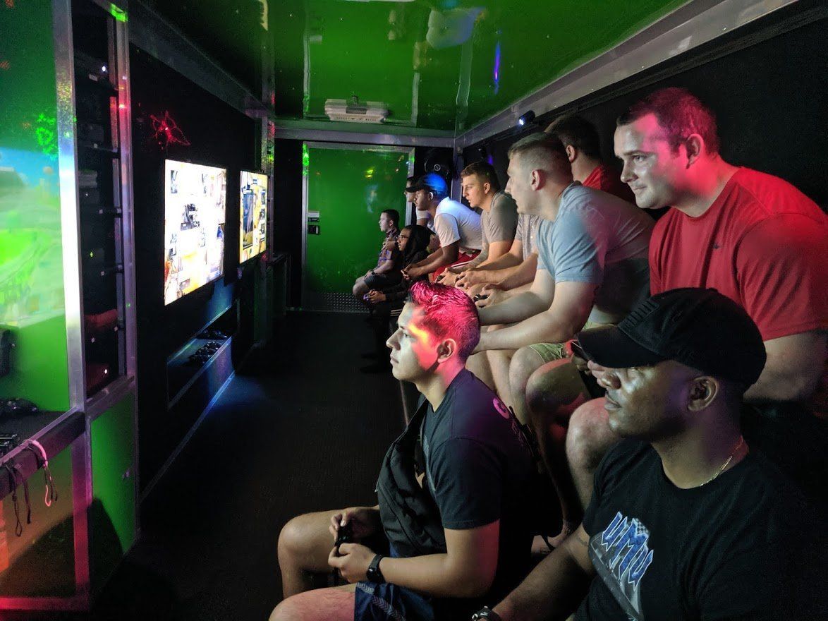 A group of people seated in a gaming trailer, intently playing video games on several wall-mounted screens.