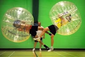 Two people wearing bubble suits collide while playing bubble soccer in an indoor court with a bright green wall.
