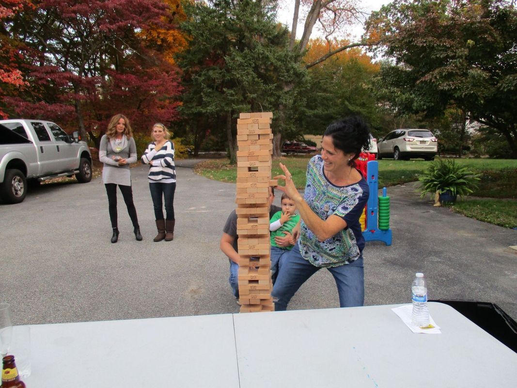 People playing a giant outdoor Jenga game on a driveway, with a table in the foreground and vehicles parked nearby.
