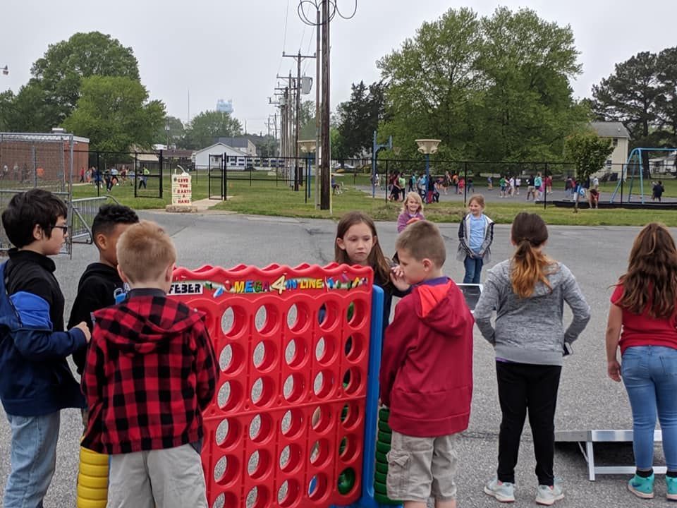 Children gather around a large, colorful Connect Four game on an outdoor school playground.