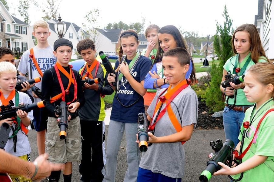 A group of people wearing orange and green gear holding laser tag equipment outside a suburban house.