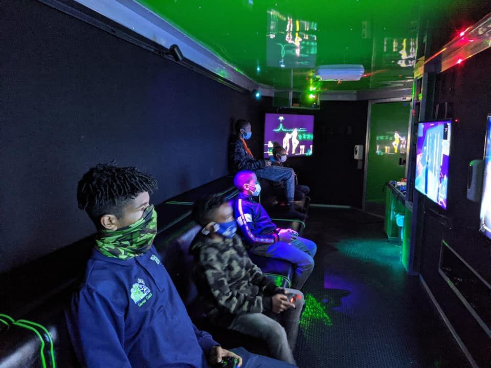 People wearing face masks sit on a bench inside a dark gaming truck playing video games on multiple wall-mounted screens.
