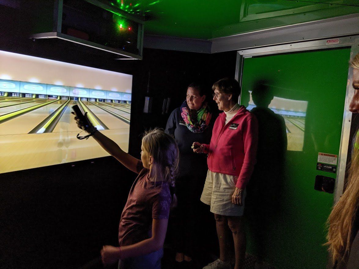A child points a remote at a large digital bowling screen, while two adults watch in a green-lit room.