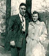 A man and woman stands outdoors by a tree, wearing coats and facing the camera.