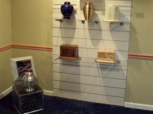 Wall-mounted display of urns and plaques in a hallway, with a framed photo and memorial items on a table