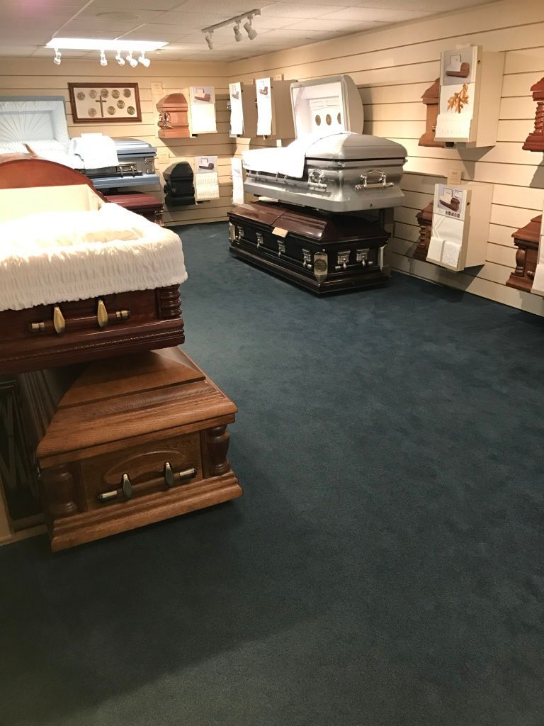 Coffins displayed in a funeral home showroom with carpeted floor and wall shelves.