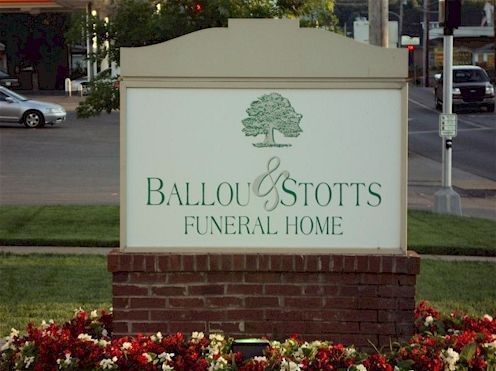 Ballou & Stotts Funeral Home sign with tree logo, set above a brick base and flower bed.