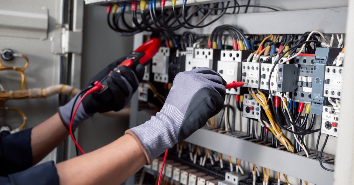 An electrician wearing safety gloves servicing a panel with various wires and connections in an industrial setting.