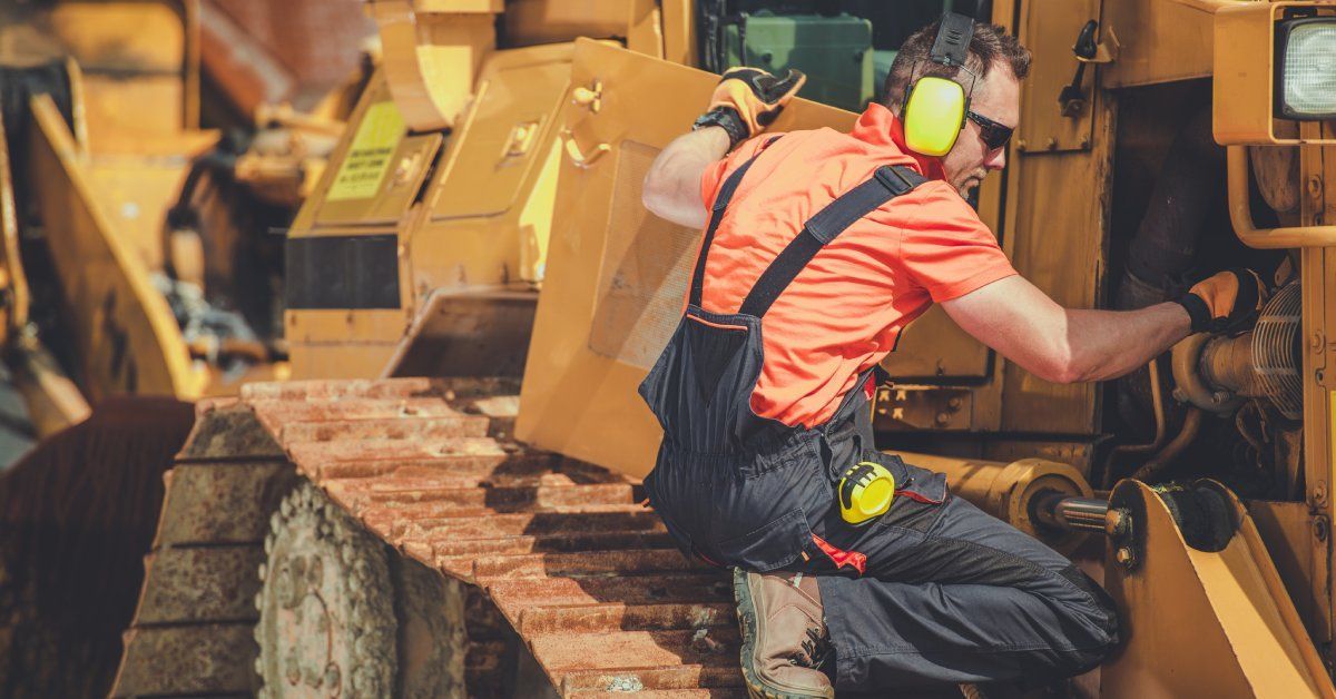 A construction worker wearing overalls and ear protection kneeling in front of heavy equipment performing repairs.