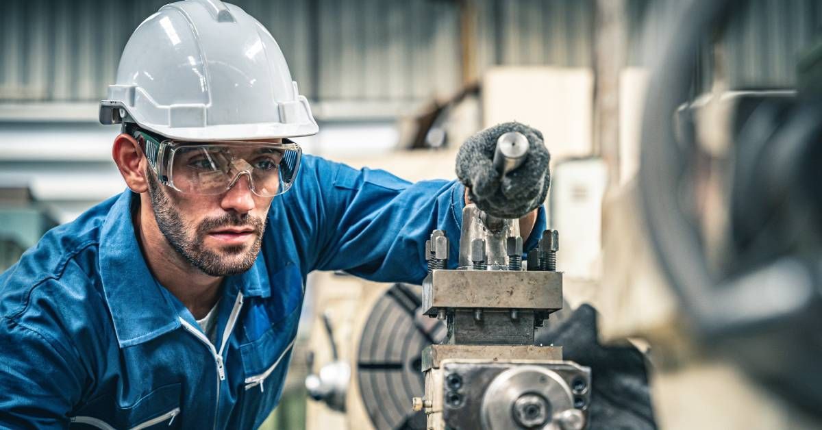 A worker wearing a blue jumpsuit and white hard hat operating a manual metal lathe machine in a factory.