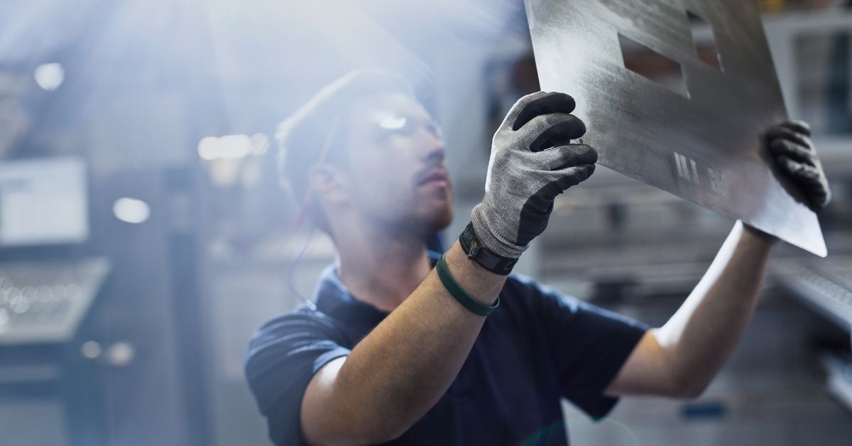 A man wearing gloves and protective eyewear holding up a piece of sheet metal with holes punched in it for inspection.