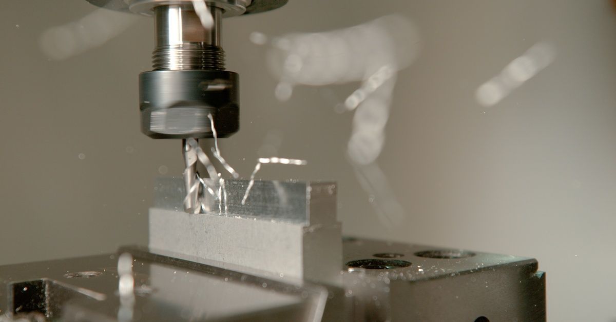 A CNC machine uses a spinning drill to remove shreds of excess material from a small stationary piece of aluminum.