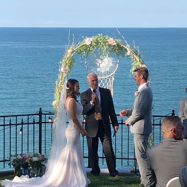 Wedding ceremony by the ocean. Bride, groom, officiant under floral arch; guests in background.