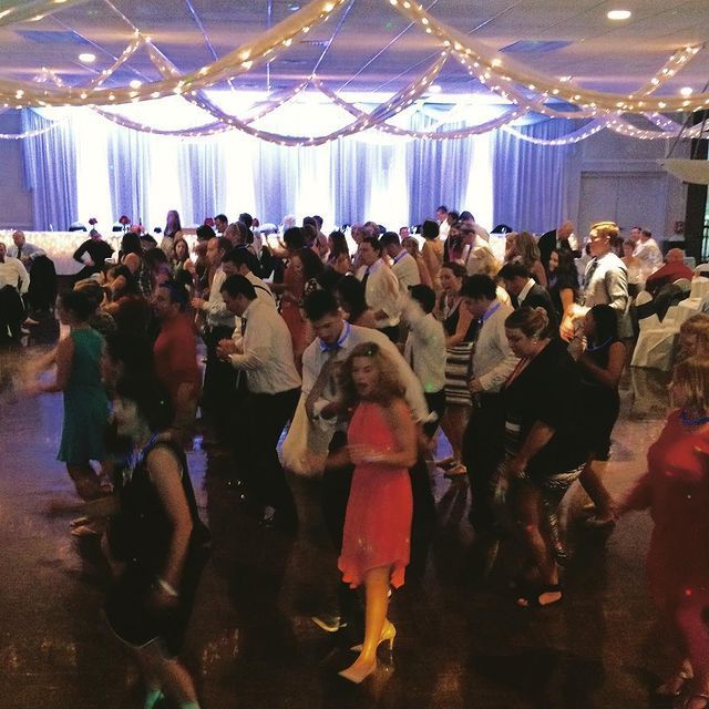 People dancing in a brightly lit ballroom. Some wearing formal attire, others in casual wear. String lights hang from the ceiling.