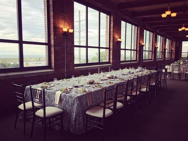 Long table set for a formal event in a room with large windows, brick walls, and decorative sconces.