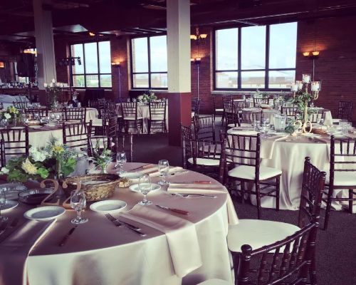 Banquet hall set for event, round tables with ivory linens, dark wood chairs, windows.