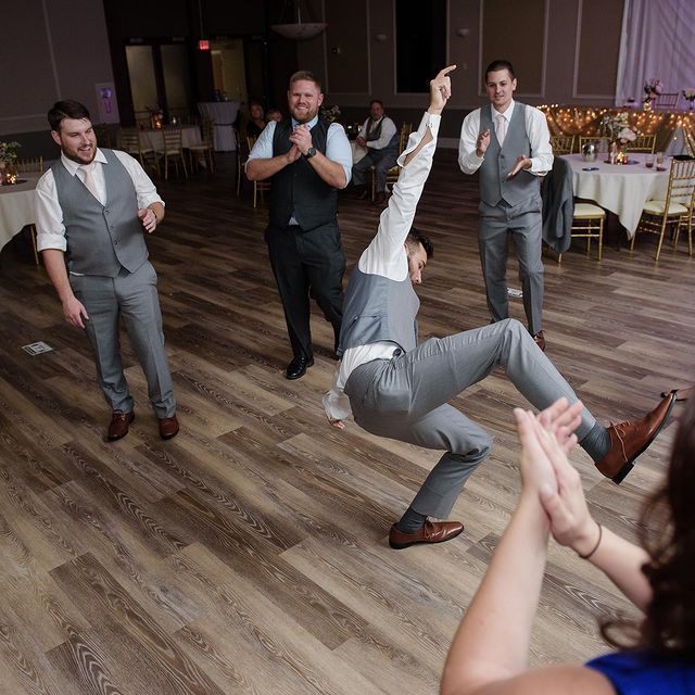Men in gray suits dancing at a reception; one person is mid-air, others clap.