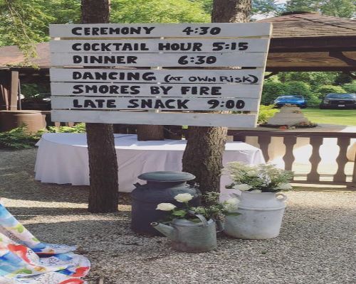 Wedding event schedule on a wooden sign attached to a tree, featuring ceremony, cocktail hour, dinner, dancing, s'mores, and late snack times.