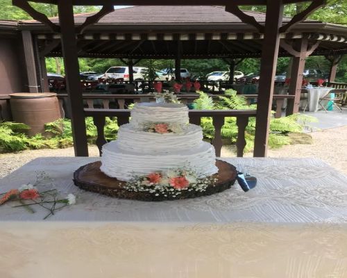 Three-tiered white wedding cake with coral flowers on a wooden slab, under a gazebo.