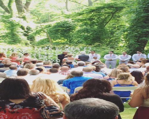 Wedding ceremony in a wooded area; guests seated, wedding party standing at altar.