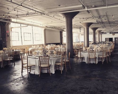 Event space with round tables, white tablecloths, gold chairs, and tall columns.