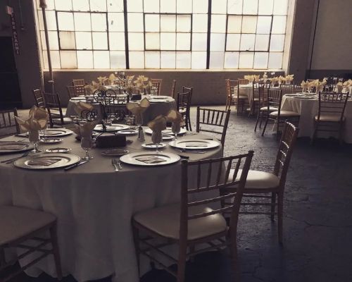 Tables set for a formal event in a room with large windows. Chairs surround tables with centerpieces.