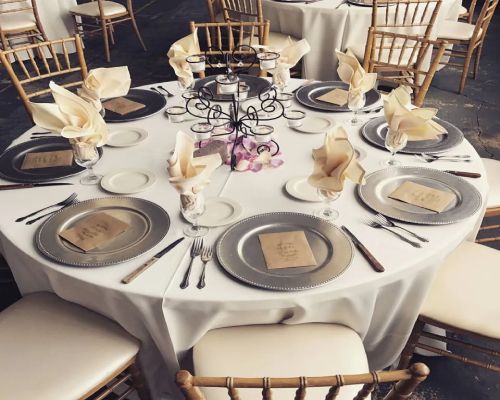 Round dining table set for a formal event with silver chargers, white plates, and beige napkins.