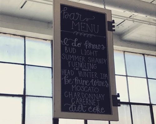 Bar menu sign hanging in front of windows, listing beers, wines, and soft drinks.