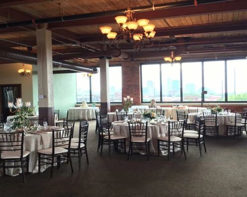 Interior of a venue with round tables set for a formal event, featuring chandeliers, large windows, and city views.
