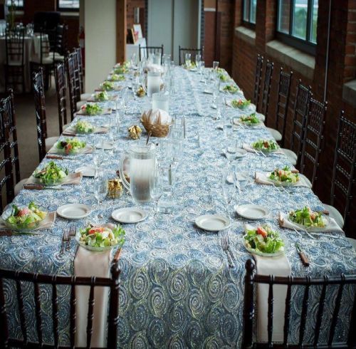 Long dining table set for a meal, blue tablecloth, salads on plates, chairs on each side, brick wall.