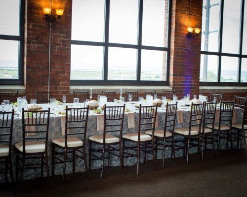 Long rectangular table set for a wedding reception in a brick-walled room with large windows, overlooking a view.